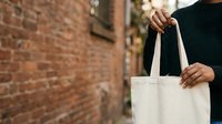 Person of mixed ethnicity wearing black sweater holding white canvas tote bag against brick wall background for eco-friendly shopping and sustainable lifestyle concepts.