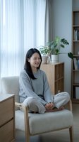 Young Asian woman in cozy gray sweater sitting cross-legged on modern armchair in bright living room with plants and natural light for wellness lifestyle.