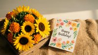 Bright sunflower bouquet with orange chrysanthemums in wooden box next to floral happy birthday greeting card on burlap surface for celebration gifts.