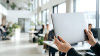 Hands holding laptop with blurred office background, colleagues working at desks. Suitable for business, technology, corporate workspace, remote work, and professional team concepts.