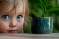 Curious young Caucasian girl with bright blue eyes and blonde hair peering at small green ceramic pot with plant sprout on wooden surface against blurred background.