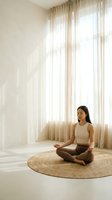 Young woman of East Asian descent in meditation pose on round jute rug, seated in lotus position wearing beige tank top and brown leggings, practicing yoga and mindfulness in minimalist bright room with sheer curtains.
