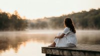 Young woman in white shirt sitting on wooden dock at peaceful lake during golden hour sunrise with misty water and forest background for wellness content.