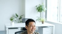 Asian male professional in casual business attire sitting at modern white desk with potted plants, bright window background, suitable for corporate, business, and workplace concepts.