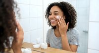 Young African American woman applying face cream in bathroom mirror wearing striped shirt during morning skincare routine for healthy glowing skin.