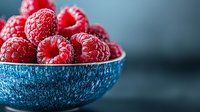 Fresh ripe raspberries in blue ceramic bowl against dark background. Macro shot with selective focus highlighting vibrant red fruit texture.