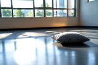 Soft gray pillow on empty studio floor with natural sunlight streaming through windows creating dramatic shadows and peaceful minimalist atmosphere.