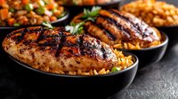 Grilled chicken breast with charred grill marks served on bed of seasoned rice in black bowl, with vegetables and herbs garnish, dark moody background.