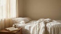 Cozy unmade bed with white linens and soft morning sunlight streaming through window curtains creating warm peaceful bedroom atmosphere.