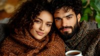 Young diverse couple in cozy knit sweaters sharing intimate moment with coffee cups, warm autumn mood, romantic lifestyle portrait against blurred background.
