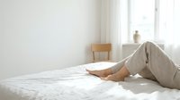 Woman relaxing on white bed in bright minimalist bedroom wearing casual beige pants. Morning comfort and peaceful lifestyle concept for wellness content.