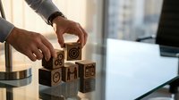 Professional businessman arranging wooden blocks with business icons on modern office desk, representing strategic planning and corporate development concepts.