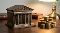 Miniature bronze bank building model with classical columns sits on wooden desk beside stacked gold coins, symbolizing financial services and banking.