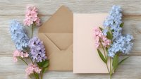 Delicate pink and blue hyacinth flowers frame kraft paper envelope and blank card on rustic wooden background, creating romantic spring composition for wedding stationery.