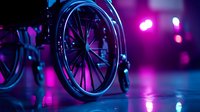 Wheelchair wheel close-up in neon lighting with purple and blue colors reflecting on floor, creating dramatic nightclub atmosphere for healthcare and accessibility concepts.