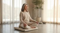 Peaceful mature woman meditating in lotus position on cushion in bright modern living room with natural light and minimalist decor for wellness.