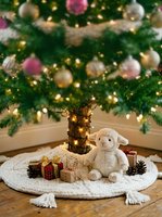 Cream-colored plush lamb toy surrounded by wrapped gifts and pinecones on white knitted tree skirt beneath decorated evergreen with gold lights and ornaments.
