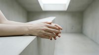 Hands clasped together in empty white room with skylight, representing solitude, meditation, prayer, mental health, isolation, and contemplation.