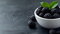 Fresh ripe blackberries in white ceramic bowl with mint leaves on dark stone background, scattered berries around, moody food photography composition.
