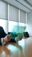 Businessman in dark suit holding transparent tablet displaying financial growth charts and data analytics with upward trending graphs in modern office setting.