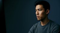 Asian male in his thirties wearing casual gray t-shirt, looking thoughtfully to the side against dark blue background. Studio portrait for business, lifestyle, and editorial use.