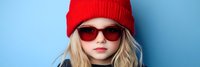 Stylish blonde child girl wearing red knit beanie hat and sunglasses against blue background. Cool kid fashion portrait for trendy clothing campaigns.