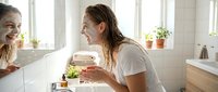 Young woman with long brown hair and fair complexion applying skincare product to her face in bright bathroom with mirror reflection, natural morning routine and beauty care.