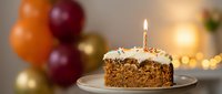 Chocolate birthday cake slice with lit candle and cream frosting on plate. Colorful balloons and warm lights create festive celebration atmosphere.