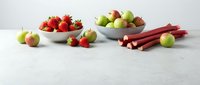 Fresh seasonal produce arrangement with strawberries, apples and rhubarb stalks in white bowls on clean marble surface for healthy cooking concepts.