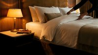 Cozy hotel bedroom with warm lighting, white bedding, wooden nightstand and glowing table lamp creating intimate hospitality atmosphere for relaxation.