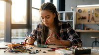 Young African American woman artist with braided hair creating detailed artwork at desk with art supplies in bright modern studio workspace.