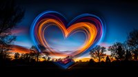 Glowing heart shape created with light painting against dramatic sunset sky and silhouetted trees, combining orange and blue colors in long exposure photography.