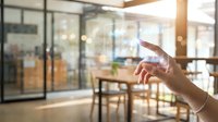 Hand holding smartphone in modern office space with glass walls and natural sunlight streaming through windows creating warm atmosphere for business communication.