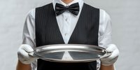 Professional waiter in formal attire with black vest, bow tie, and white gloves holding empty silver serving tray against neutral background.