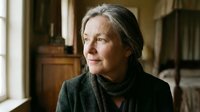 Mature Caucasian woman with gray hair in dark sweater sitting thoughtfully by window in cozy home interior for lifestyle and aging concepts.