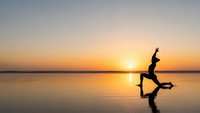 Woman practicing yoga warrior pose silhouette at sunrise over calm water with golden sky reflection for wellness and meditation concepts.