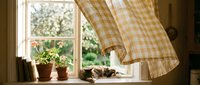 Young woman with fair skin and blonde hair arranging potted plants on a sunny windowsill with gingham curtains, creating a cozy cottage-style home decor scene.