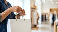 Close-up of woman's hands holding white shopping bag in modern retail store with blurred clothing racks and warm lighting in background.