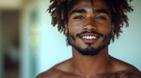 Young African American man with dreadlocks smiling warmly at camera showing genuine happiness and positive energy in bright indoor setting.