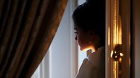 Young Asian woman silhouetted against window light, standing thoughtfully near elegant curtains in morning atmosphere, creating dramatic contrast and peaceful solitary moment.