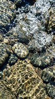 Crystal clear water flowing over smooth river rocks with sunlight creating dancing light patterns and reflections on stone surface for nature backgrounds.