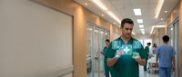Male doctor in green scrubs using digital tablet with holographic medical interface in modern hospital corridor with colleagues in background.
