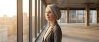Confident senior Caucasian businesswoman with gray hair stands by office windows, golden sunlight illuminating her thoughtful expression and city view.