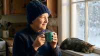 Elderly woman in knit hat enjoying hot beverage by window on snowy winter day with sleeping cat nearby creating cozy domestic scene for seasonal lifestyle content.