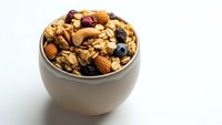 Homemade granola with nuts, dried fruits and seeds in ceramic bowl on white background. Healthy breakfast cereal mix for morning meal preparation.