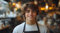 Young male barista with tousled brown hair smiling warmly in professional kitchen wearing apron, hospitality worker portrait for restaurant industry.