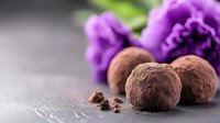 Dark chocolate truffles with cocoa powder coating on dark background with purple flowers, selective focus on confectionery with blurred floral decoration.