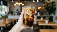 Unrecognizable person holding cold cola drink with ice in cafe setting with bokeh lights and blurred interior background.