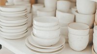 Handmade ceramic bowls and plates in natural beige tones stacked on wooden surface, artisan pottery collection for kitchen and dining room decor.