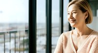 Middle-aged Asian woman in casual pink cardigan gazing out from high-rise window overlooking cityscape, expressing contentment and reflection for lifestyle and wellness concepts.